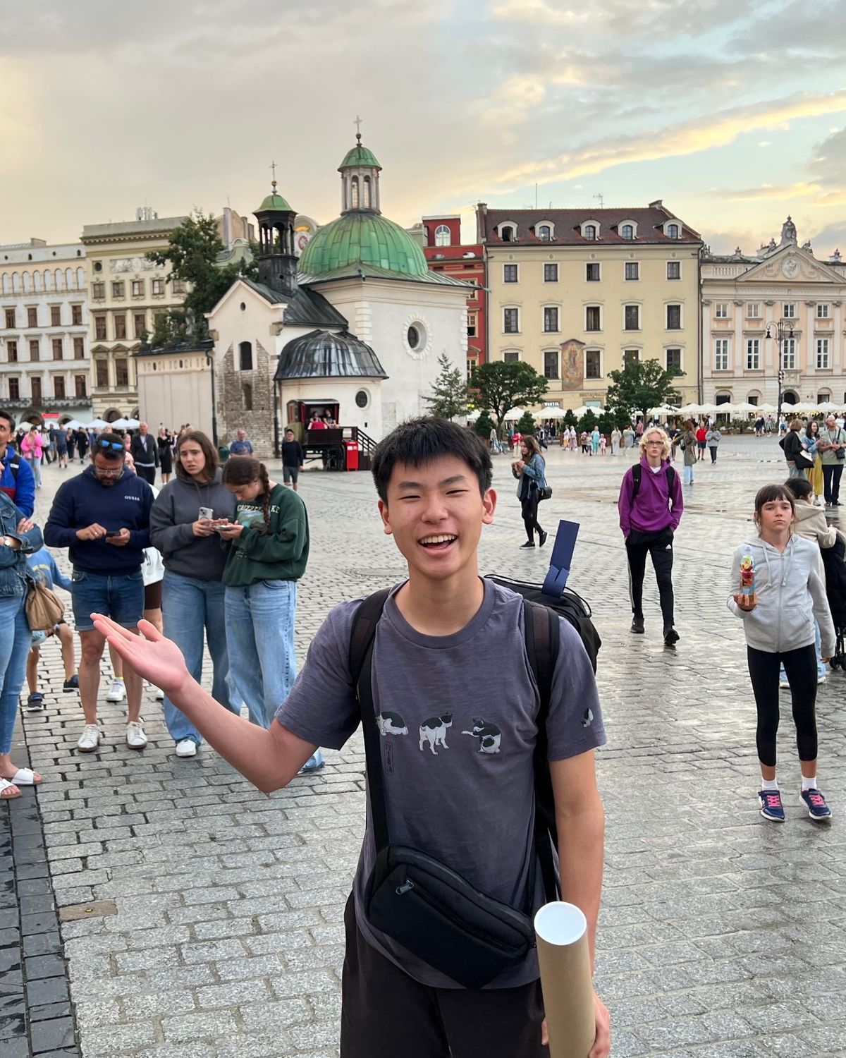 Ethan laughing and gesturing toward Kraków's Main Market Square at sunset, with the green-domed Church of St. Adalbert behind him