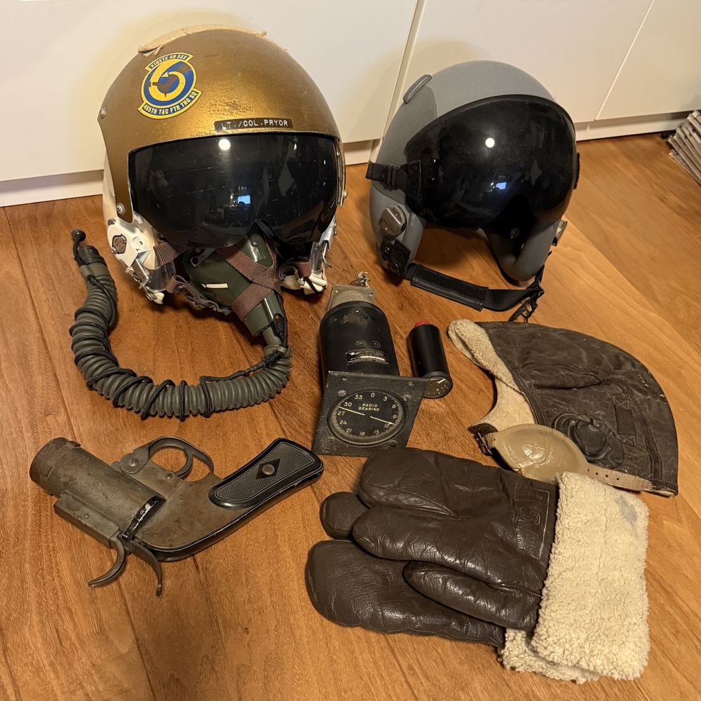 Two pilot's flight helmets with visors and oxygen masks, a flare pistol, an aircraft gauge, a leather flight cap, and sheepskin-lined gloves