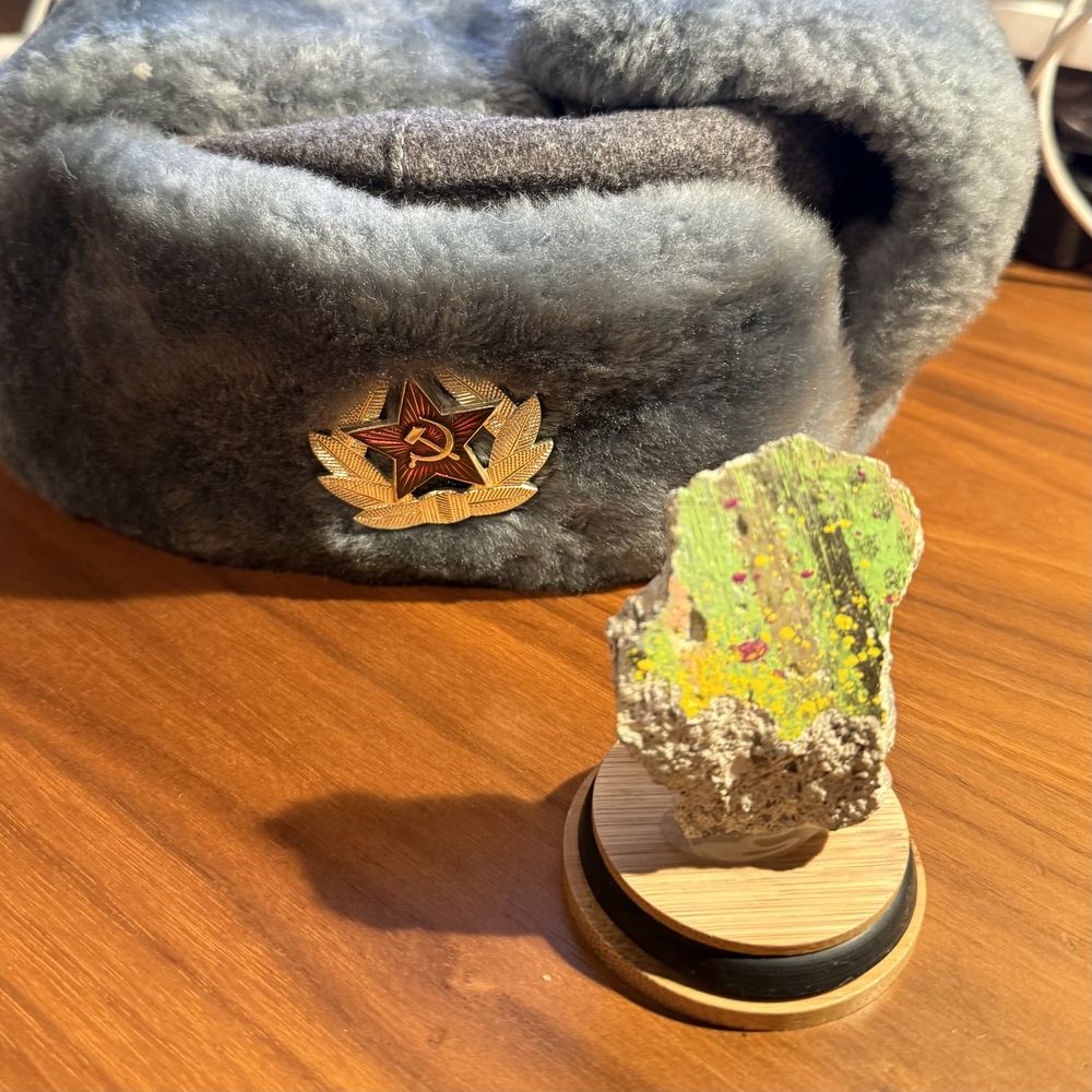 A painted fragment of the Berlin Wall on a wooden stand in front of a Soviet fur hat with a hammer-and-sickle badge