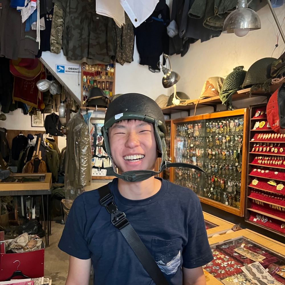 Ethan laughing while trying on an old military helmet in a crowded militaria shop full of uniforms, medals, and badges