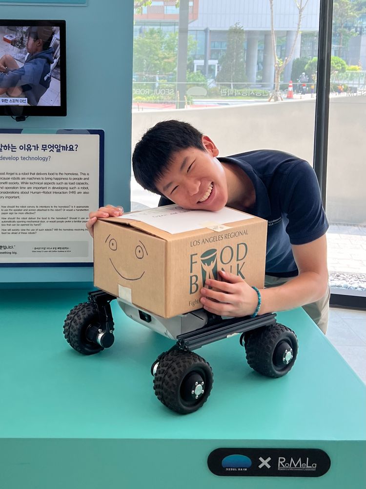 Ethan hugging Food Angel, his cardboard-box robot with a drawn smiley face, on permanent display at the Seoul Robot & AI Museum