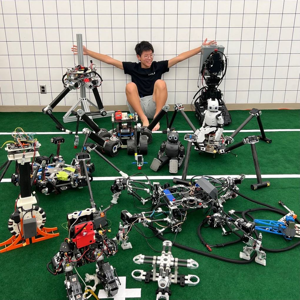 Ethan sitting against the wall of the RoMeLa lab with his arms spread wide, surrounded by more than a dozen robots