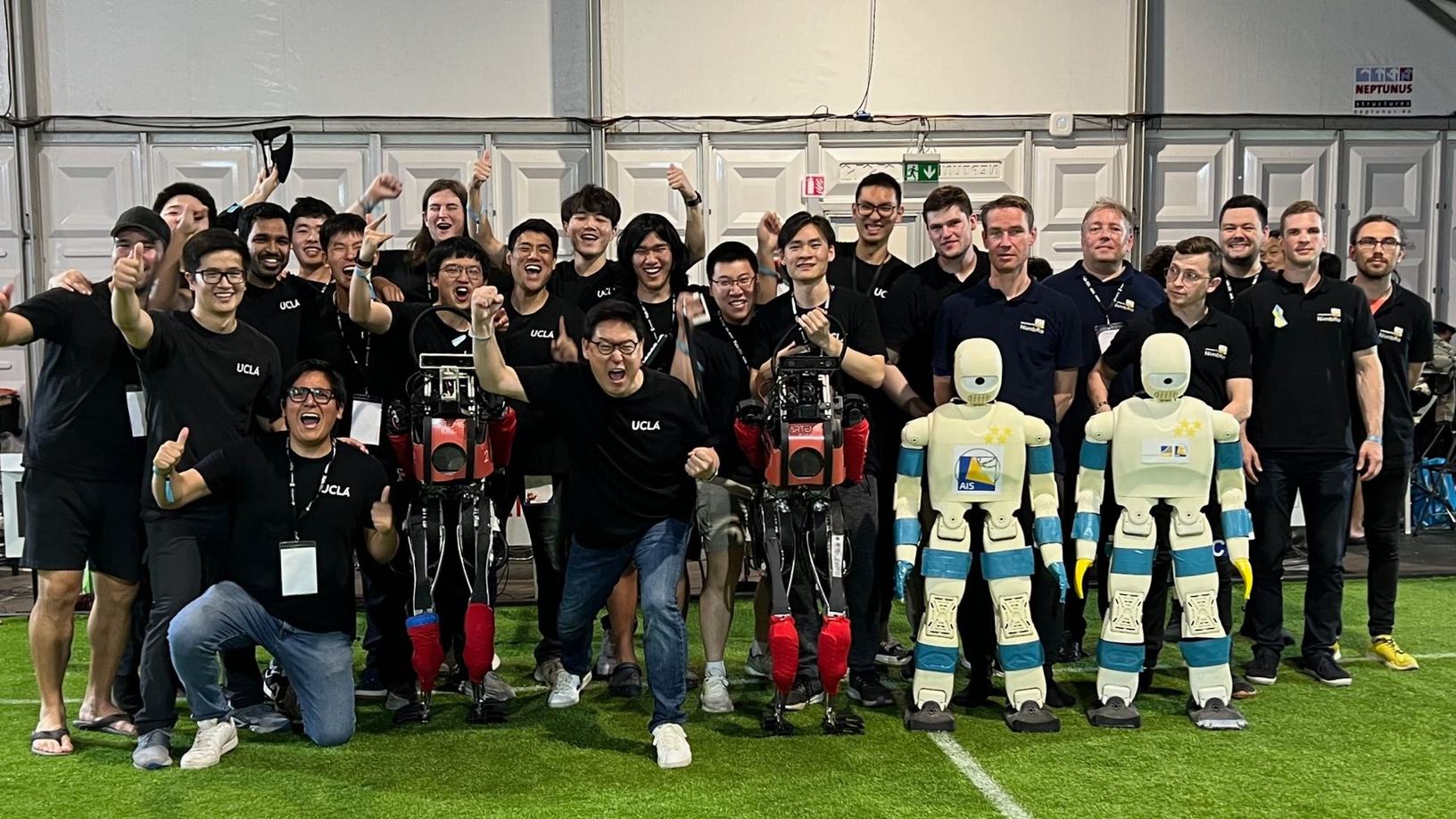 Team RoMeLa from UCLA and Team NimbRo from the University of Bonn posing together on the field with their humanoid robots