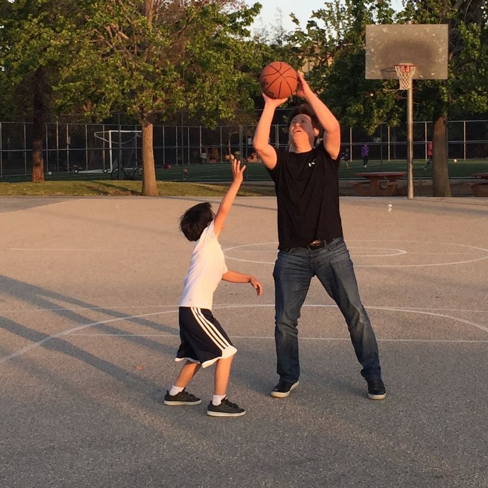 Young Ethan reaching up to block a much taller player's shot on a park court at sunset