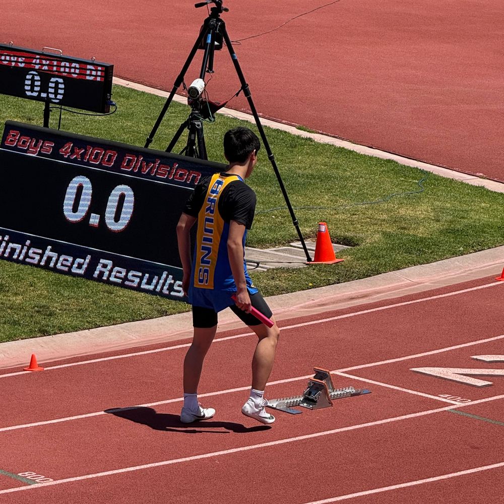 Ethan from behind in a Bruins singlet, holding a relay baton beside the starting blocks and a Boys 4x100 Division results board